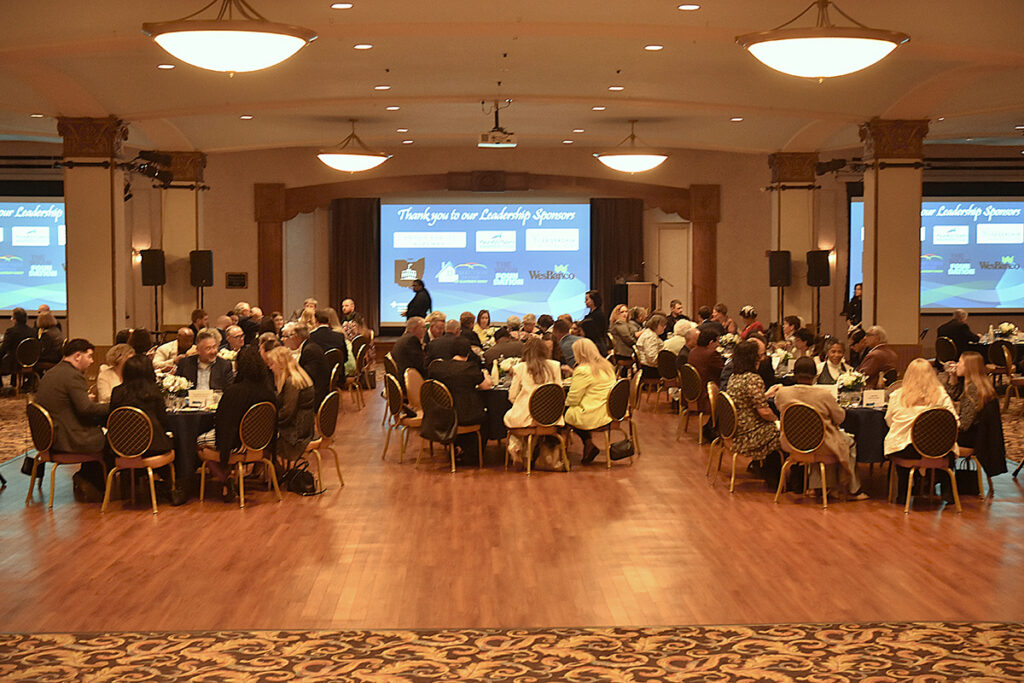 A large banquet hall with several round tables, each hosting formally dressed attendees engaged in conversation. Large screens display sponsor logos at the front. The atmosphere is elegant and professional.