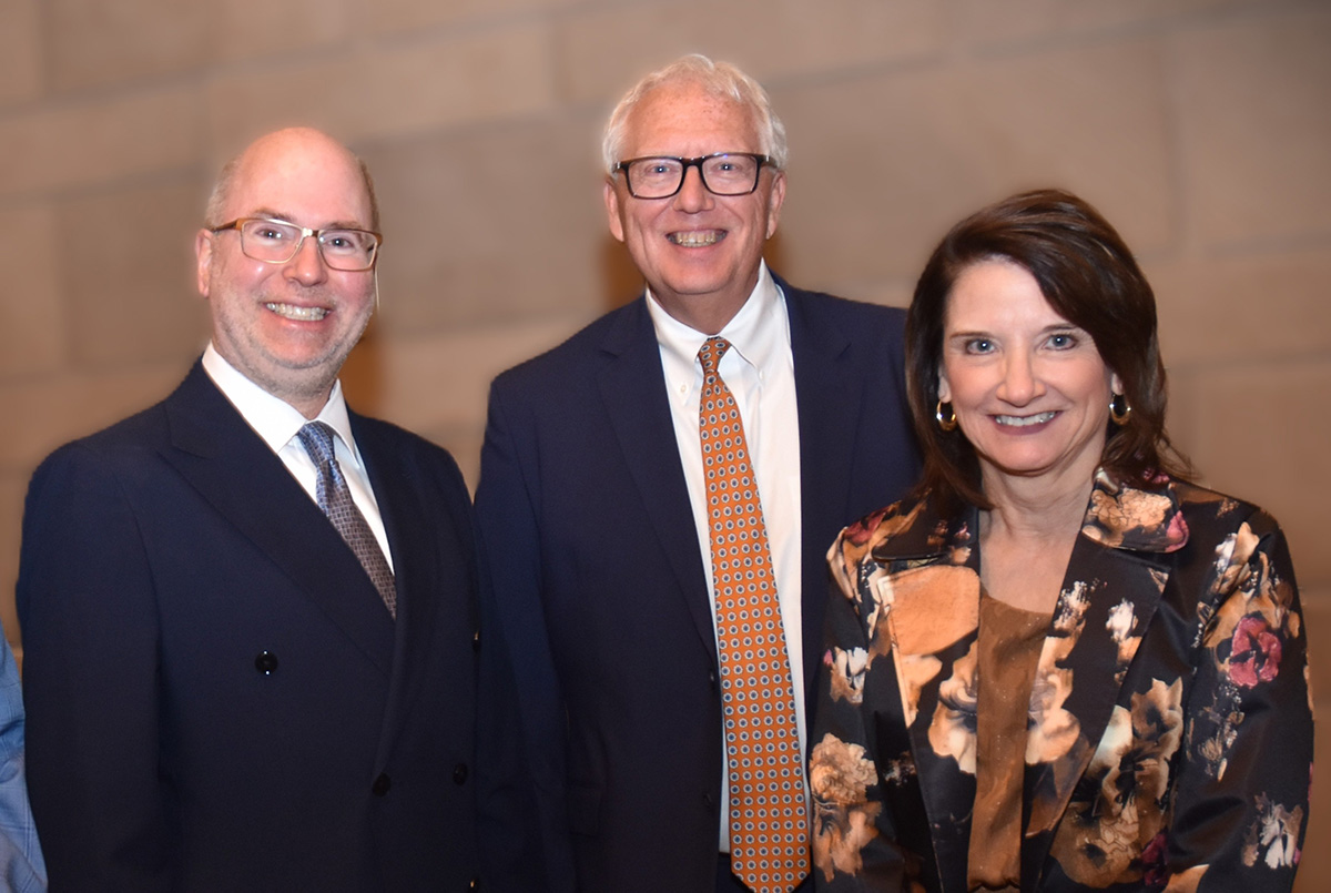 Three people, two men and a woman, pose together smiling in formal attire. They stand in front of a neutral-colored wall, conveying a cheerful tone.