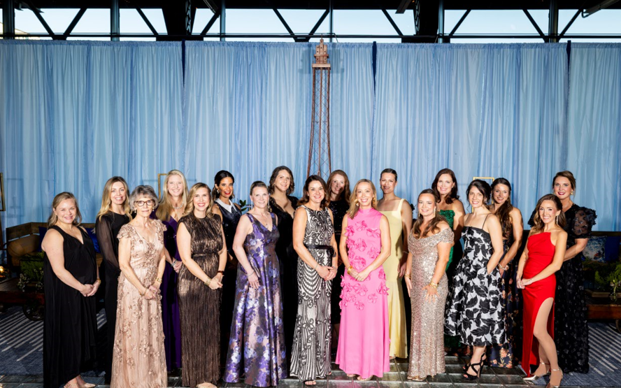 A group of women in elegant evening gowns stand in a row, smiling. The background features soft blue drapes, conveying a formal and celebratory atmosphere.