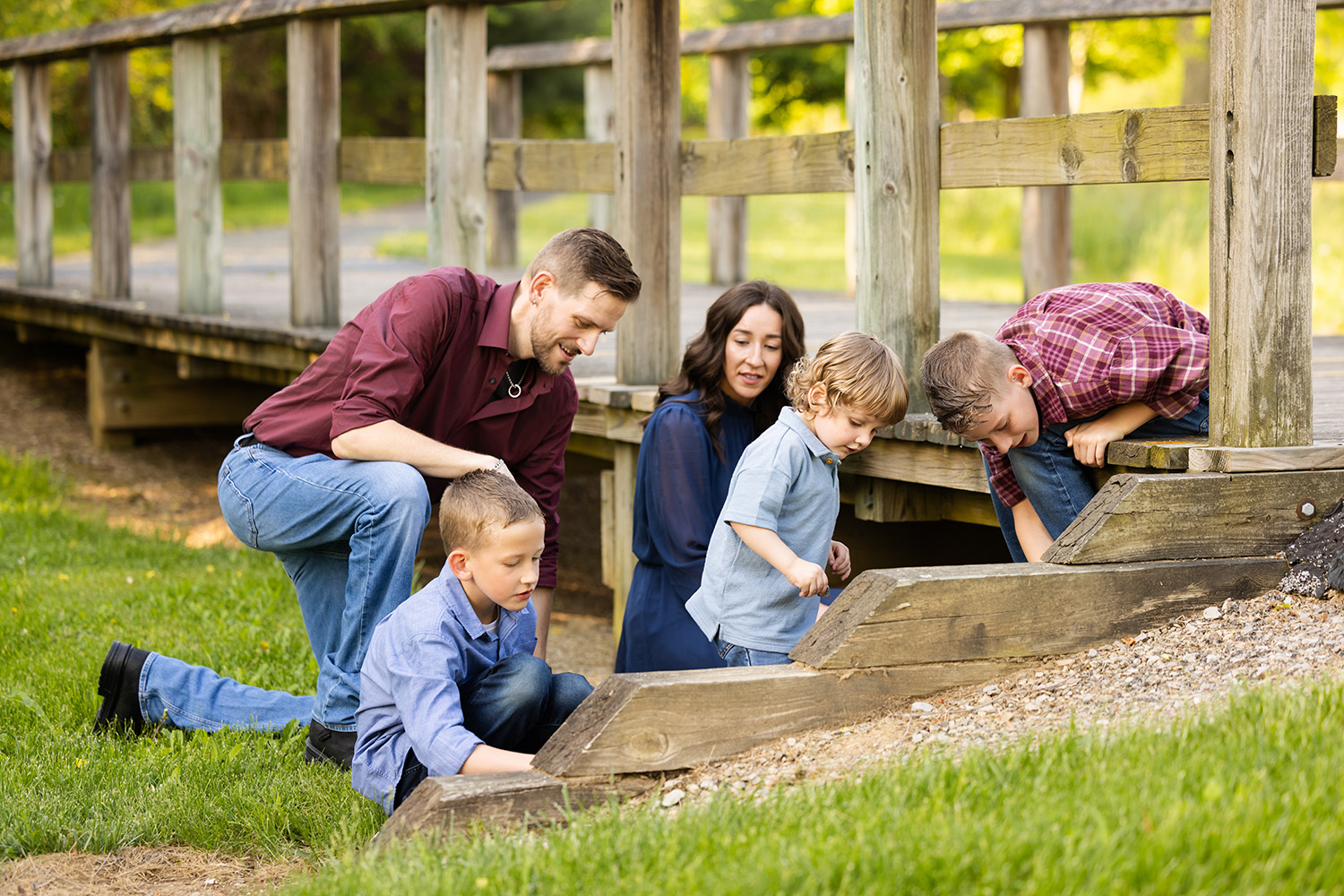 A family of five crouches near a wooden bridge, exploring the ground with curiosity. The setting is bright and green, conveying a sense of warmth and togetherness.