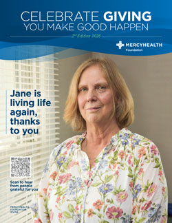 Portrait of a smiling woman in a floral blouse, standing in front of a Merseyhealth Foundation donor banner with the text 'Jane is living life again, thanks to you' and a QR code.