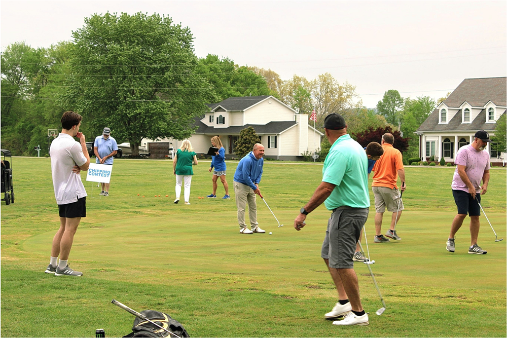 Group of people on a grassy field playing golf, a 'Chipping Contest' sign visible in the background.