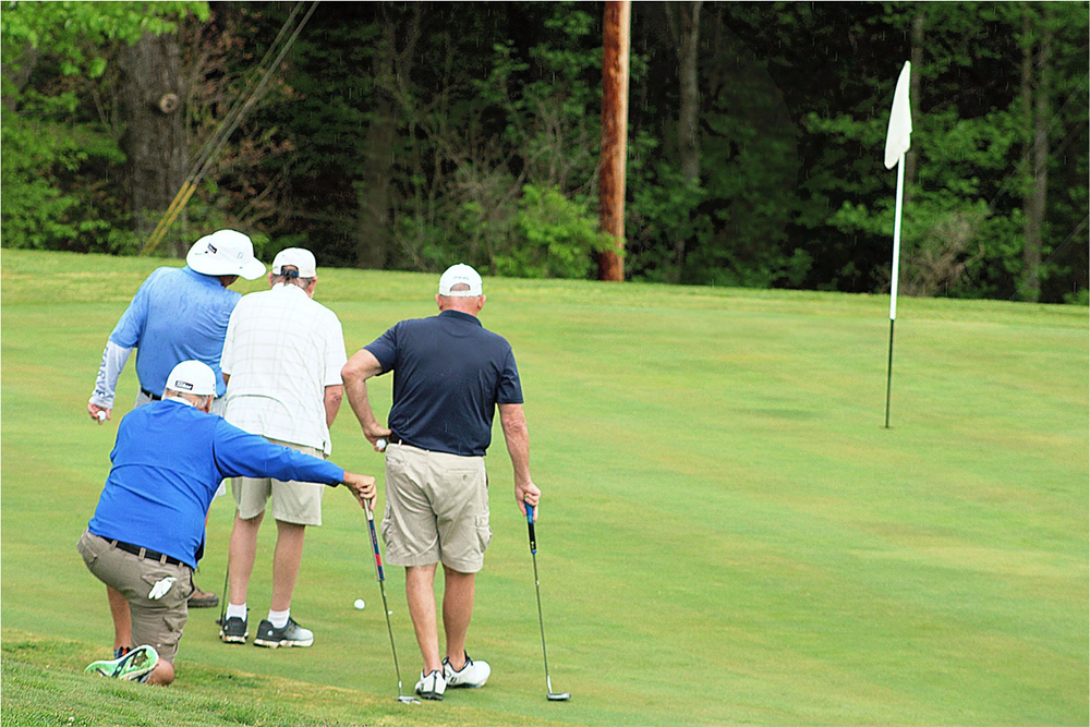 Four golfers line up a putt on a green near the hole, with one kneeling to align their shot.