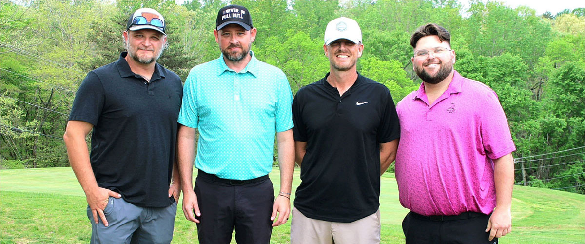 Four men pose together on a golf course, wearing polo shirts and caps, smiling at the camera on a sunny day.