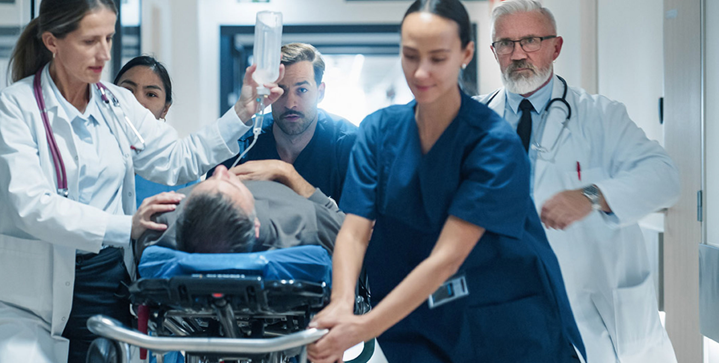 Emergency room scene with clinicians moving a patient on a stretcher, one holding an IV bag as others assist.