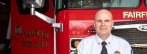 Firefighter in dress uniform standing in front of a red Fairfield Fire Department fire truck, smiling at the camera.