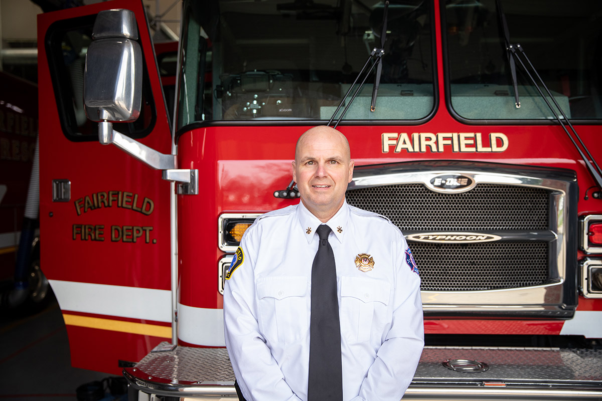 Firefighter in uniform standing in front of a red Fairfield Fire Department truck with chrome grille and badges visible.