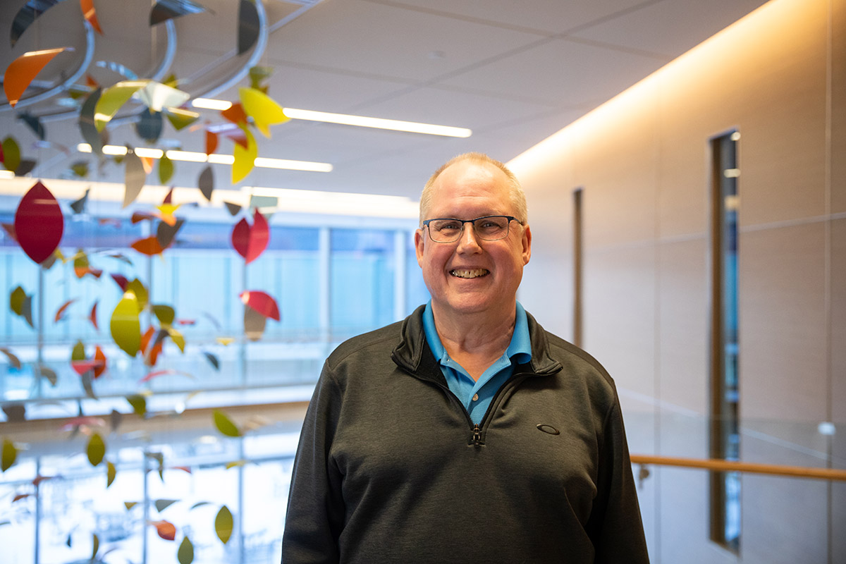 Smiling man with glasses stands in a modern indoor atrium with colorful hanging leaf decorations behind him.