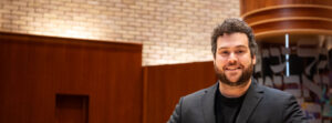 Smiling man with dark curly hair and beard in a dark blazer standing in a wood-paneled interior.