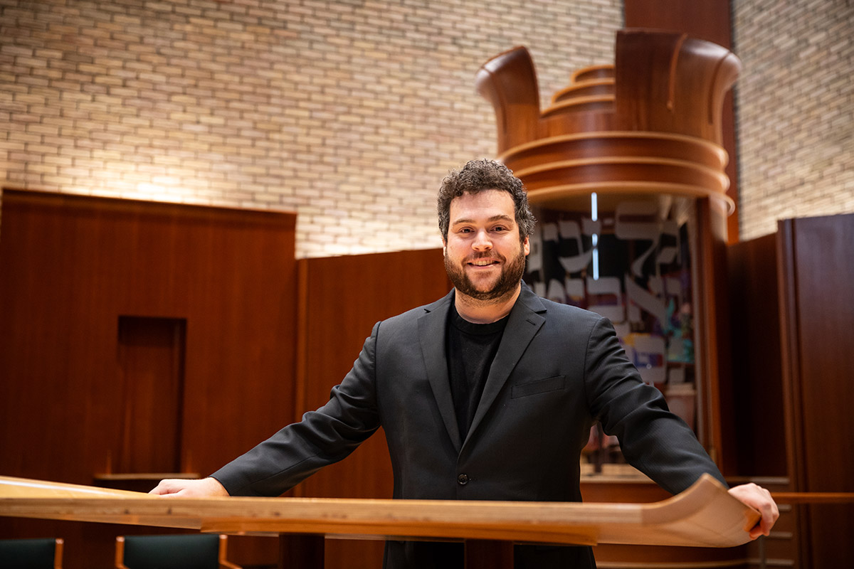 Smiling man in a dark suit standing behind a wooden railing in a warm, wood-paneled room with a large sculptural wood backdrop behind him.
