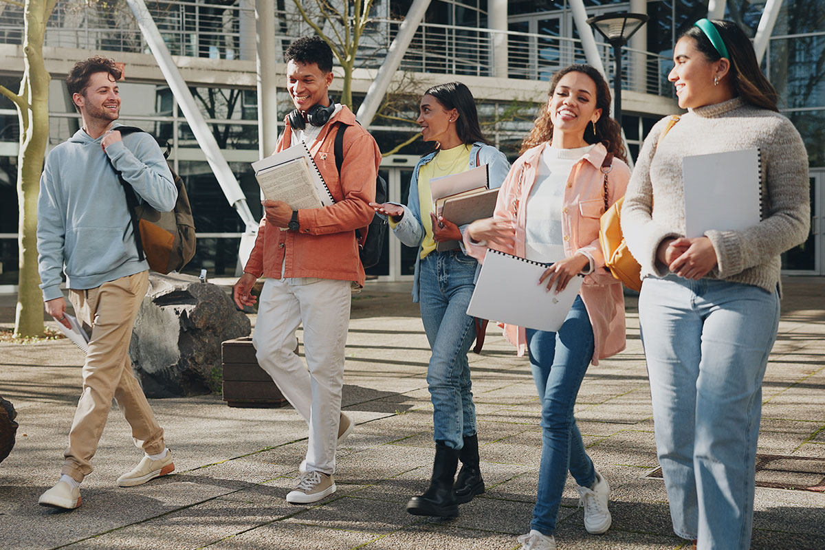 Group of diverse students walking outdoors on campus, carrying notebooks and backpacks, smiling and chatting.