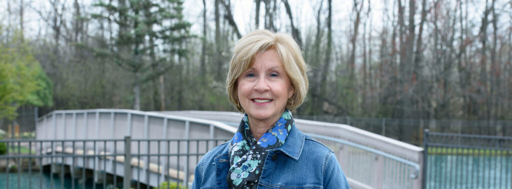 Smiling woman in a denim jacket and floral scarf stands outdoors by a railing over water and a bridge in the background.