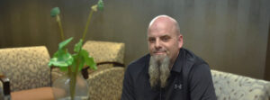 Man with a long gray beard and shaved head sits in a lounge, smiling slightly, with patterned chairs and a plant in the background.]","Bald man with a goatee sits in a beige-patterned seating area, wearing a black polo and smiling.
