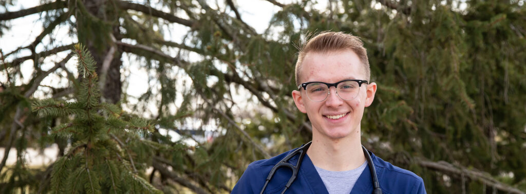 Young medical professional in blue scrubs with a stethoscope outdoors, standing in front of evergreen branches.