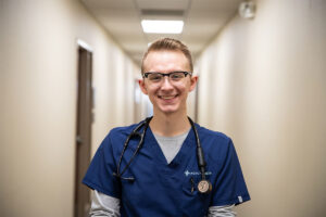 Smiling healthcare worker in blue scrubs with a stethoscope in a hospital hallway.