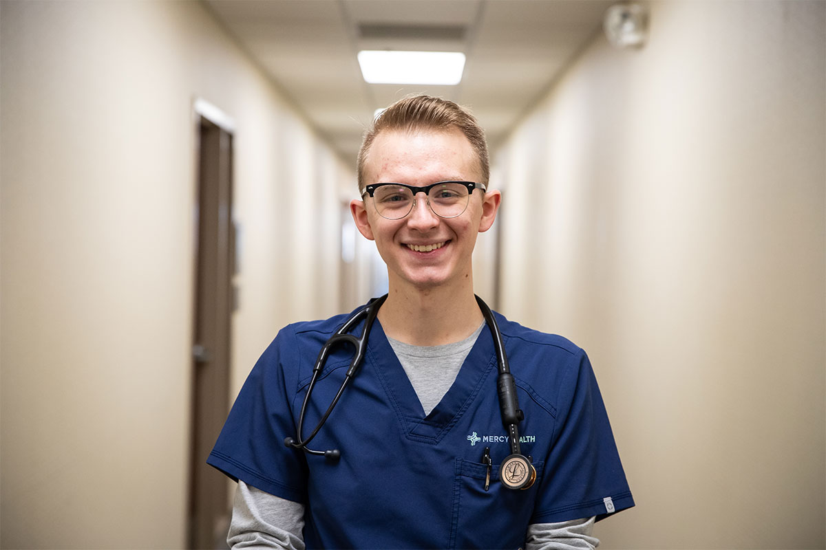 Smiling healthcare worker in blue scrubs with a stethoscope in a hospital hallway.