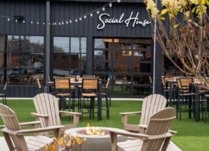 Outdoor patio of a restaurant named 'Social House' with string lights, black framed windows, and wooden tables and chairs around a fire pit.