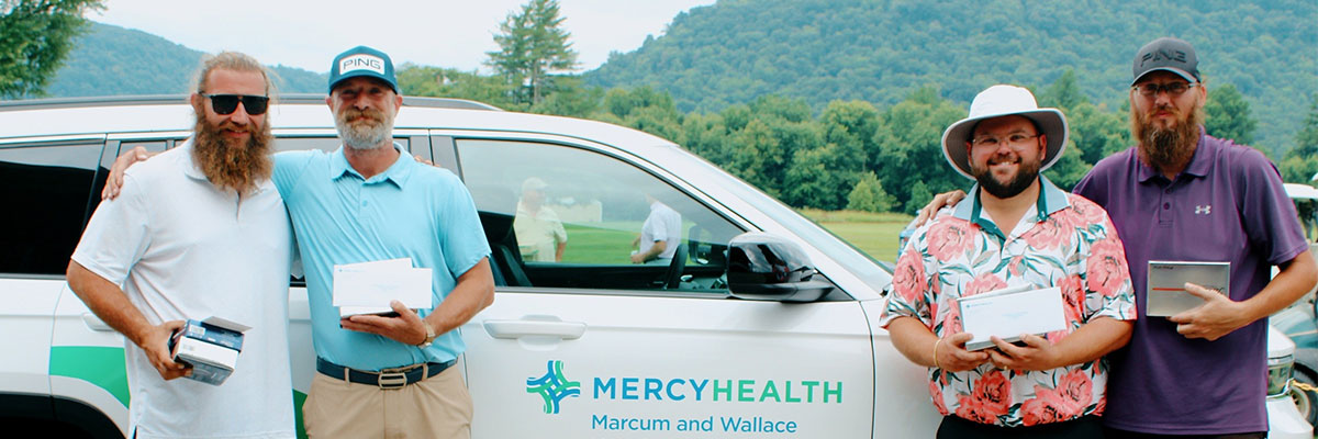 Four men pose in front of a white Mercy Health SUV with the logo, holding envelopes and small boxes outdoors.