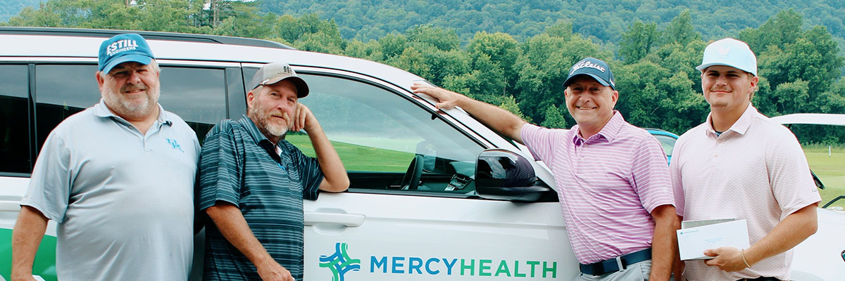 Four men stand beside a Mercy Health vehicle on a sunny day, posing for a group photo on a grassy field.