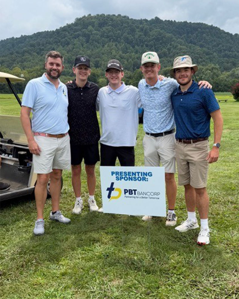 Five men in golf attire pose together on a sunny golf course, standing beside a sponsor sign that reads 'PBT Bancorp'.
