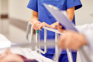 Nurse in blue scrubs pushing a hospital bed, holding a clipboard beside a patient.