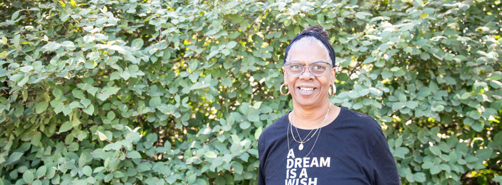 Cheerful woman wearing glasses and a head wrap stands outdoors in front of a dense green hedge, wearing a black shirt with the slogan 'A DREAM IS A WISH'.
