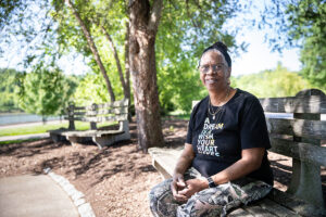 Smiling woman wearing glasses sits on a wooden park bench under leafy trees, looking at the camera.