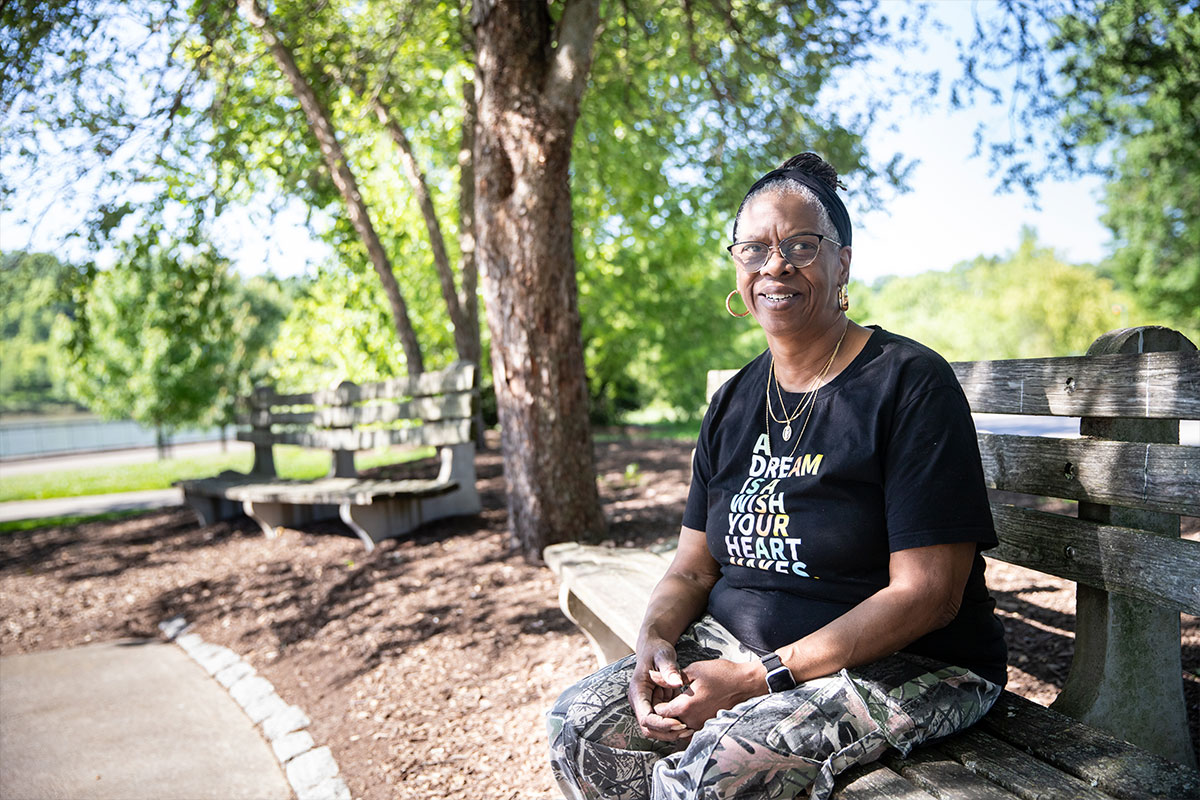 Smiling woman wearing glasses sits on a wooden park bench under leafy trees, looking at the camera.