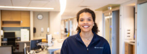 Smiling female healthcare worker in a Mercy Health uniform inside a clinic lobby or reception area.