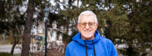 Smiling older man with gray hair and glasses wearing a blue hoodie outdoors in a park.