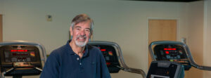 Smiling older man in a navy polo stands in a gym with treadmills behind him on a cardio room floor.