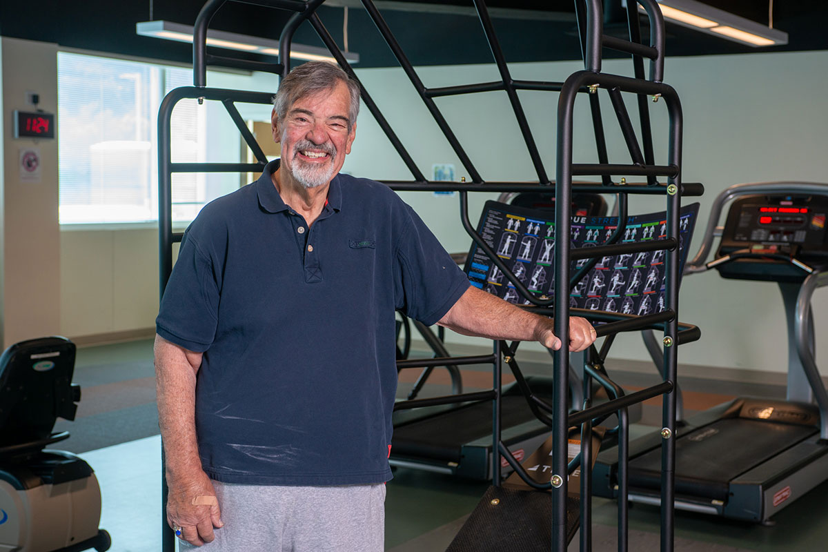 Smiling older man in a navy polo stands beside a metal exercise frame in a gym, touching the frame.