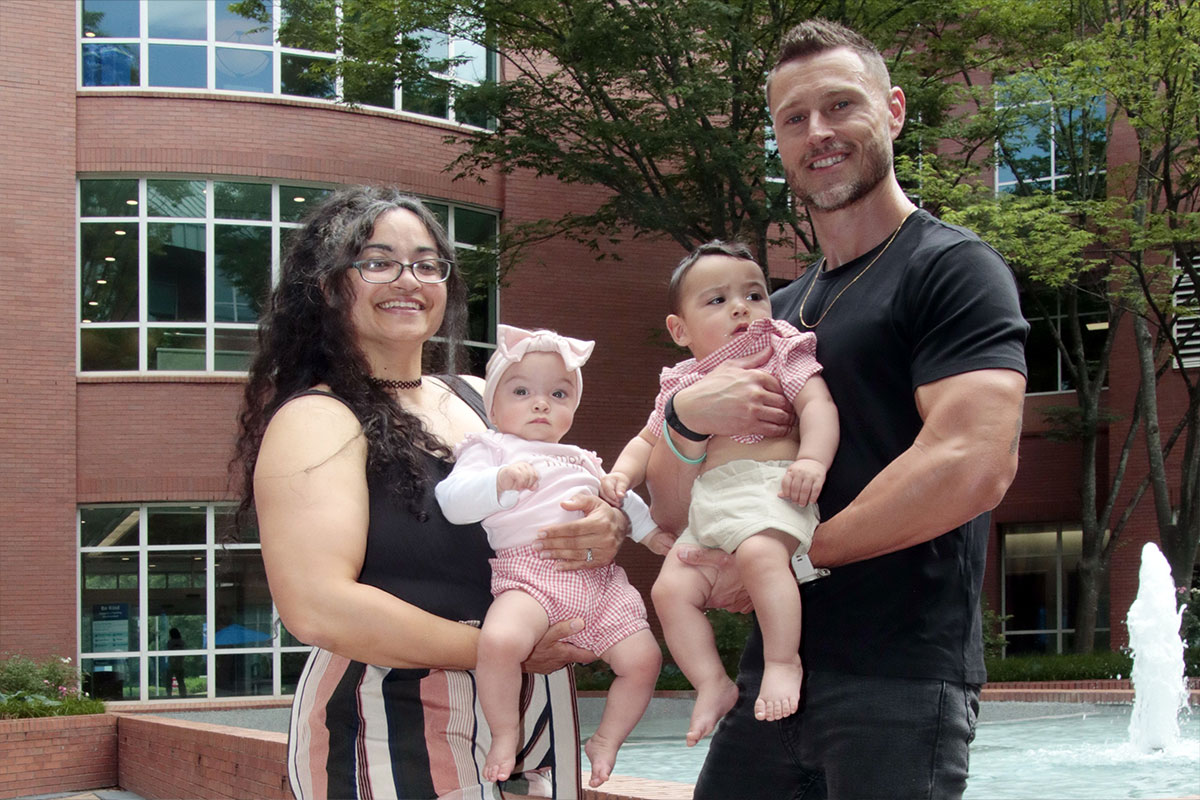 Mother and father pose with their two babies outside a brick building with a fountain in the background, smiling at the camera.