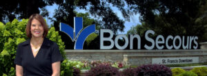 Smiling woman in a black outfit standing beside a large Bon Secours St. Francis Downtown sign outdoors.