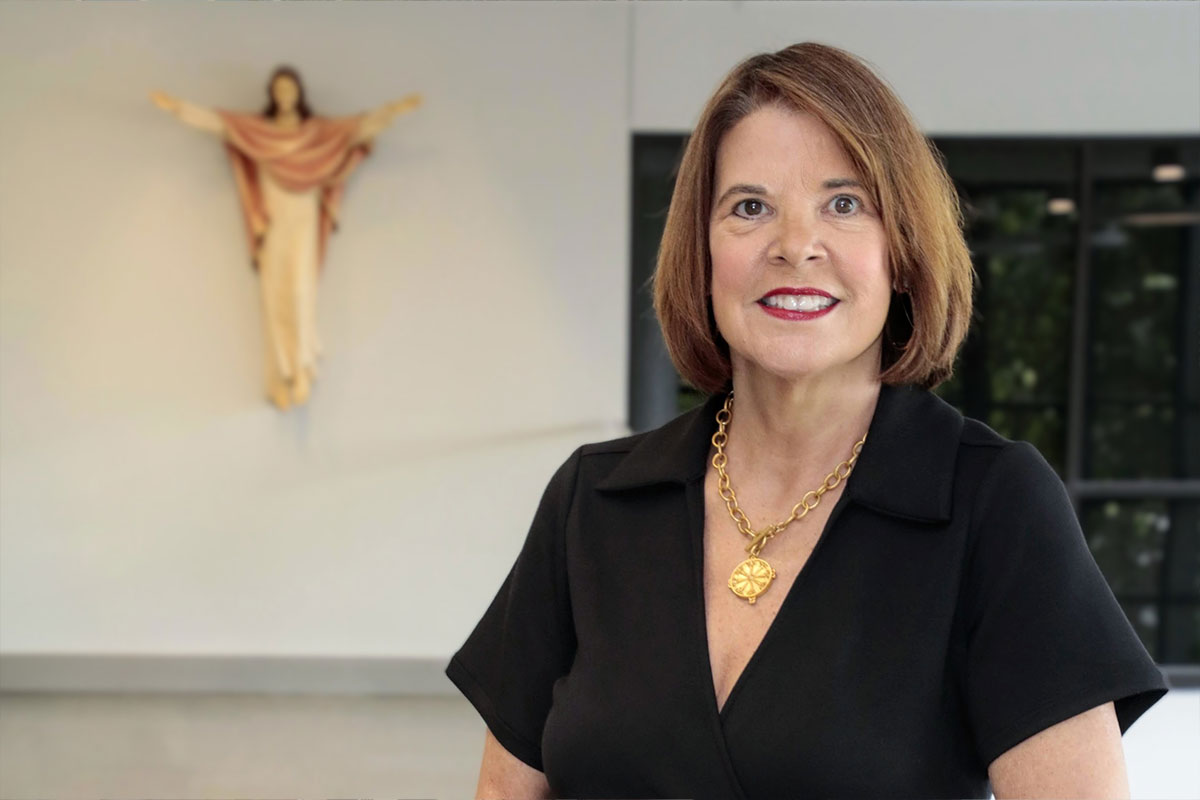 Smiling woman in a black V-neck top with a chunky gold necklace, standing in a bright interior with a crucifix on the wall behind her.