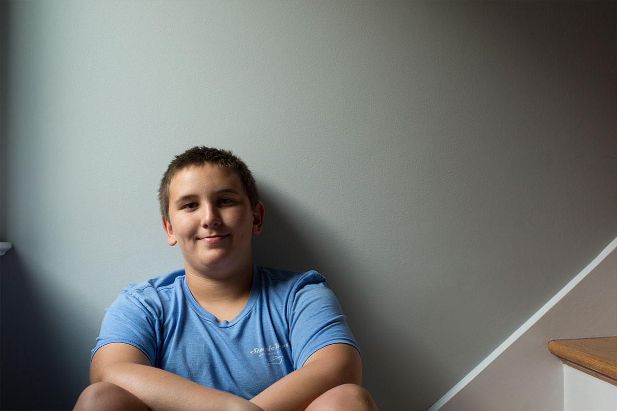 Smiling boy in a blue t-shirt sitting against a gray wall by a staircase.