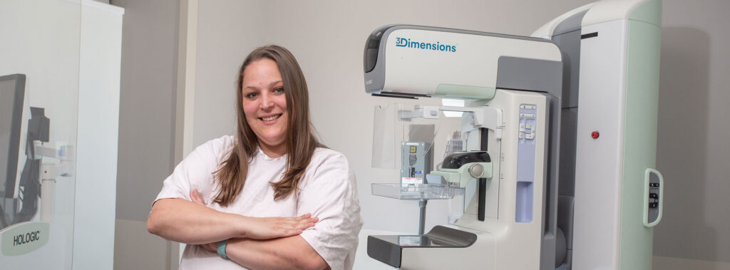 Radiographer posing beside a large 3Dimensions mammography unit in a clinical imaging room.