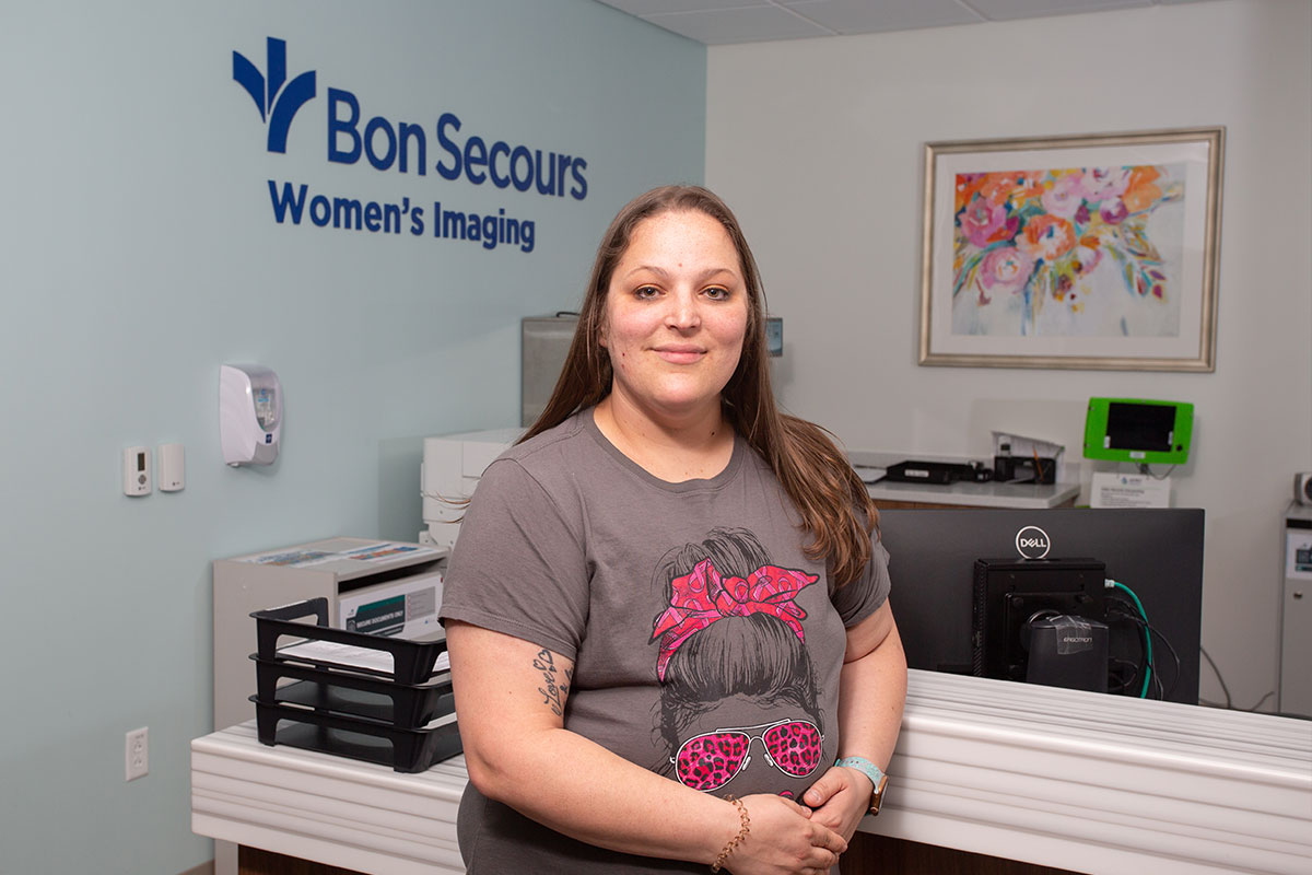 Receptionist standing at the front desk of Bon Secours Women's Imaging, smiling at the camera with the clinic sign in the background on a pale blue wall.