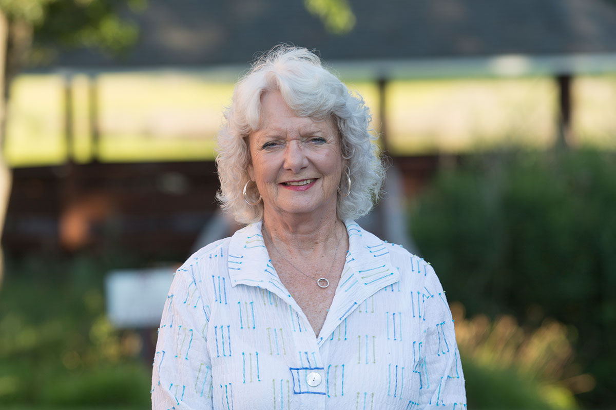 Smiling elderly woman with gray hair outdoors in a garden, wearing a white patterned blouse and hoop earrings.