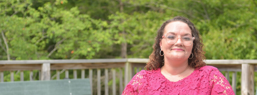 Smiling woman with glasses and a pink lace top outdoors on a wooden deck with green foliage behind her.