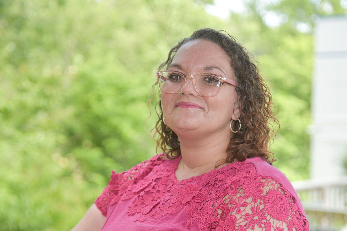 Portrait of a woman with curly hair and pink lace top, standing outdoors and smiling at the camera, with greenery in the background.