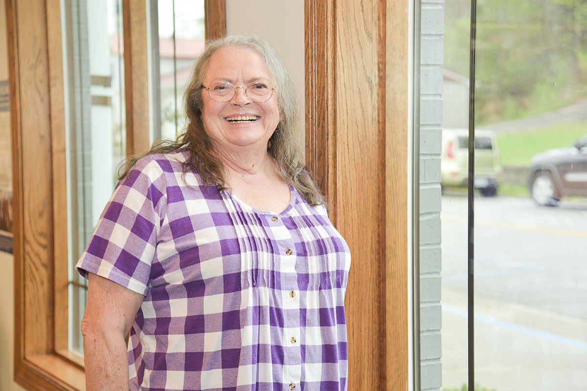 Smiling older woman with gray hair and glasses wearing a purple plaid blouse, standing by a window.
