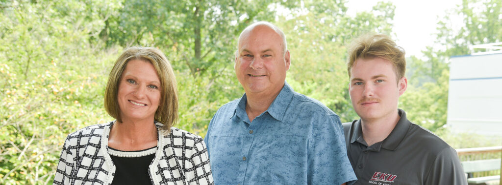 Three adults smiling and posing outdoors with greenery in the background.