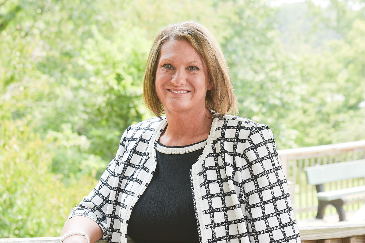 Smiling woman seated on a wooden deck with green trees in the background, wearing a black top and a white grid-pattern jacket.