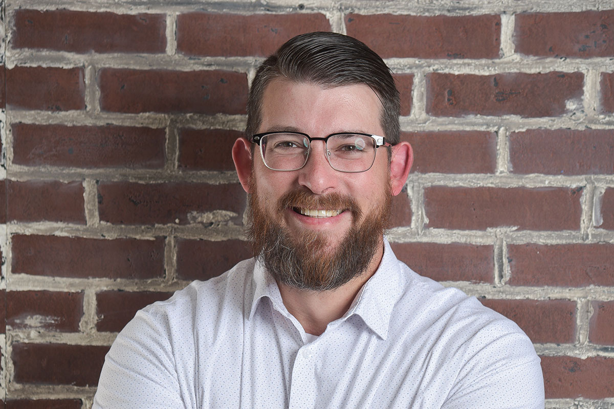 Portrait of a smiling man with a beard and glasses, wearing a light shirt, in front of a brick wall.
