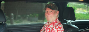 Older man with a beard wearing a red Hawaiian shirt and camo cap sits in the back seat of a car.