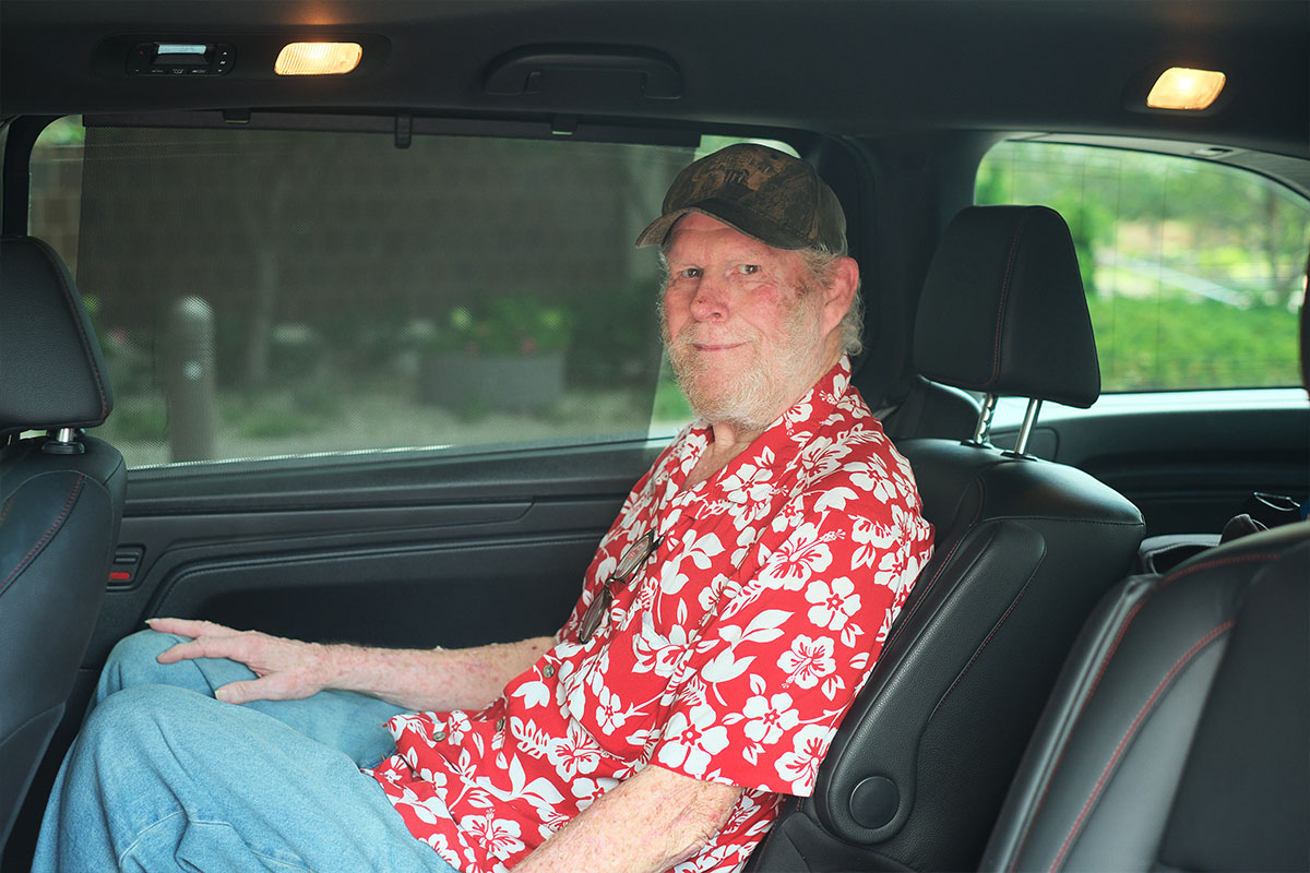 Older man with a white beard in a red floral shirt sits in the back seat of a car, looking at the camera.