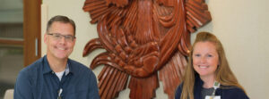 Two people pose and smile in an indoor space, with a large wooden sculpture behind them.