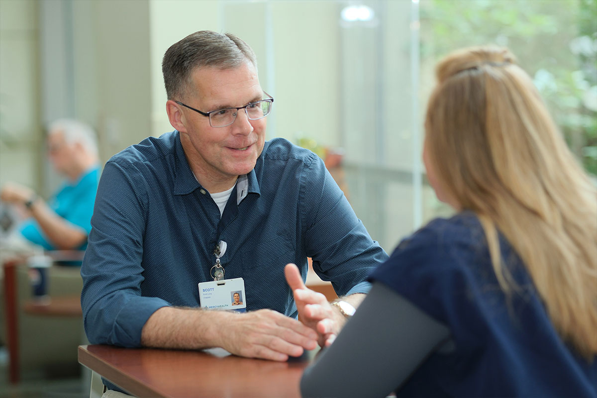 Man in a blue shirt with a name badge talks to a woman across a wooden table in a bright lobby/waiting area.