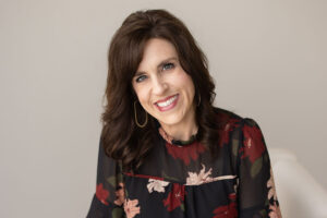 Smiling woman with shoulder-length brown hair and gold hoop earrings wearing a dark floral blouse.